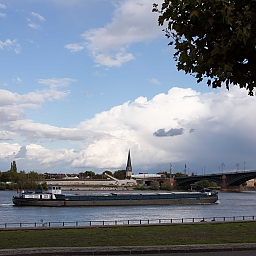 Auf dem Rhein fährt ein Lastkahn. Am anderen Ufer sieht man den Ort Kastel. Am blauen Himmel ziehen dunkle Gewitterwolken auf.
