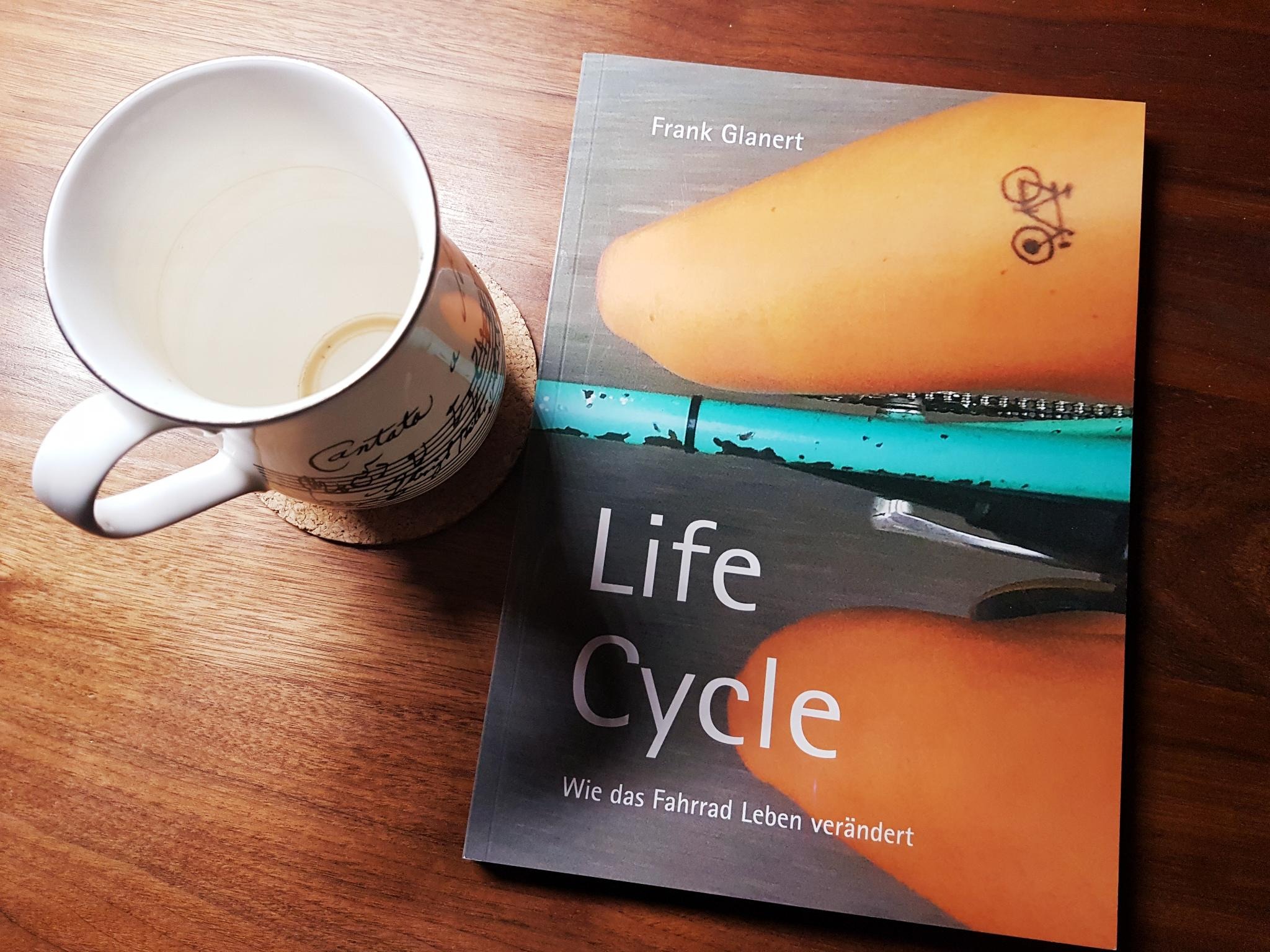 Ausgelesen: Life Cycle – Wie das Fahrrad Leben verändert. Auf einem Holztisch liegt das genannte Buch. Daneben eine leere Tasse Tee.. Bild: copy Franziska Köppe | madiko