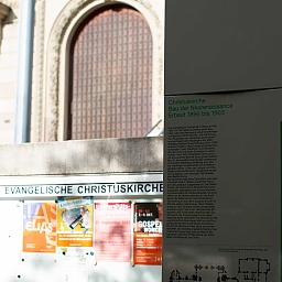 Im Vordergrund: Stehle mit Informationen zur Kirche. Im Hintergrund Aushang der Kirche und Kirchen-Hauptportal.