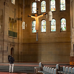 In einem schlichten Kirchenraum hängt zirka auf 6 bis 8 Metern Höhe das Kruzifix vor hellen, schlichten Kirchenfenstern.