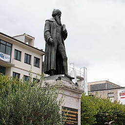 Denkmal für Gutenberg (stehend mit einem Buch in der linken Hand). Him Hintergrund ragt an einer Hauswand ein großes Transparent, das noch Werbung für die Ausstellung 