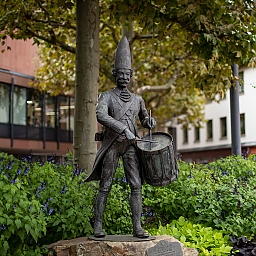 Bronzestatue eines gut gelaunten Mannes in der Tracht der Mainzer Prinzen-Garde, am Becken seitlich hängt seine Trommel, die er spielt.