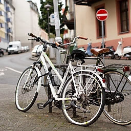Zwei Räder (eines weiß, Damenrad – eines schwarz, Herrenrad) sind gemeinsam an einen Fahrradbügel angeschlossen. Das Damenrad hat vorn und hinten einen Platten. Das Herrenrad im Retro-Stil wirkt liebevoll gepflegt mit seinem Ledersattel und Leder-Griffen.