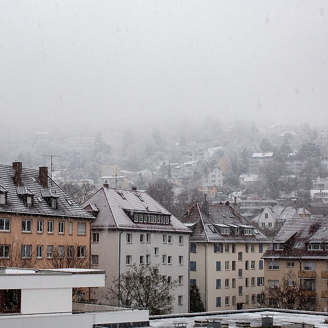 Bildbeschreibung: Halbhöhenlage im Süden des Stuttgarter Kessels. Auf den Dächern der Altbau-Gebäude liegt der erste Schnee. Schwere, große Schneeflocken fallen. Der Kesselrand ist wolken-/nebel-verhangen. Er geht direkt über in den Himmel, der mit seinem Grau-Weiß kaum Tageslicht durchlässt.