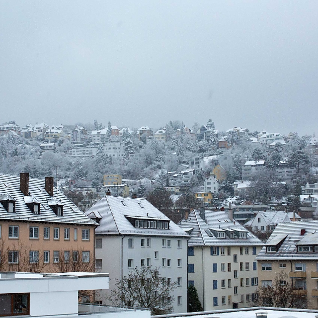 Halbhöhenlage im Süden des Stuttgarter Kessels. Über den Dächern der Altbau-Gebäude liegt der erste Schnee. Sanft schweben Schneeflocken vorbei. Der Kesselrand ist wolken-/nebel-verhangen. Er geht direkt über in den Himmel, der mit seinem Grau-Weiß kaum Tageslicht durchlässt.
