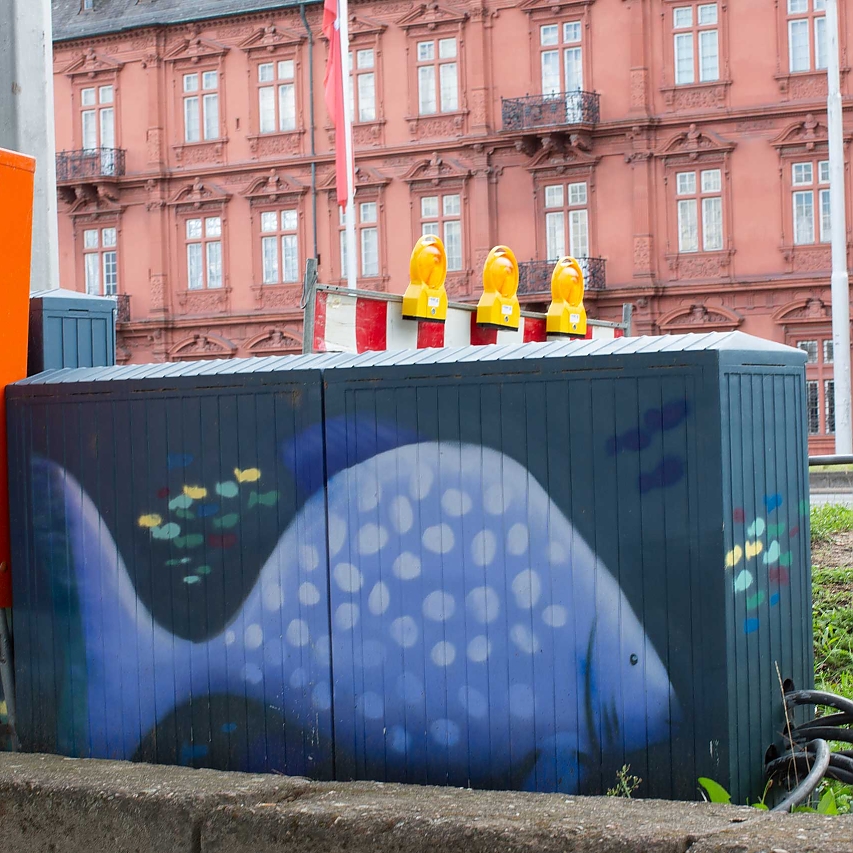 Verteilerkasten am Straßenrand geschmückt mit einem Graffiti: Blauer Fisch mit einem kleinen, bunten Fischschwarm auf dunkelgrünem Hintergrund. Im Hintergrund des Fotos sieht man den roten sandstein-Bau des Kurfürstlichen Schlosses.