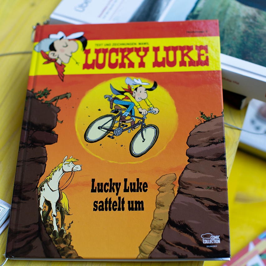 Knall-gelber Tisch. Darauf liegen verschiedene Bücher zum Thema Rad-Fahren. Im Fokus: Lucky Luke sattelt um