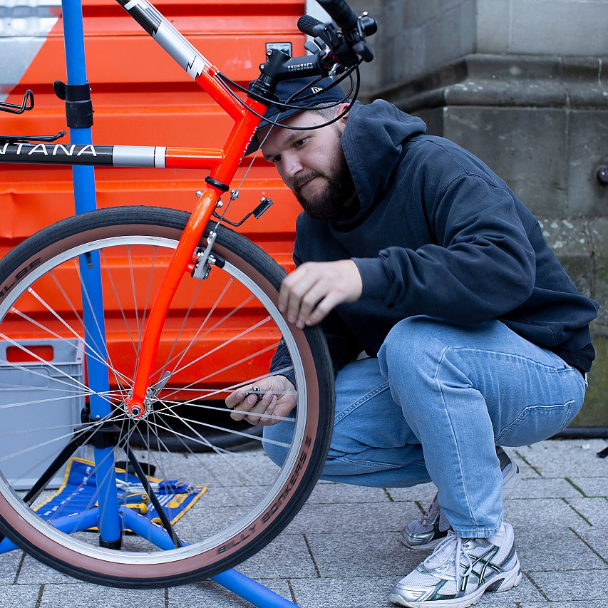 Ein Fahrrad zur Reparatur bei Lichter & Lenker: Ein Mann prüft den Reifen, Speichen und die Bremsen. Um bequemer arbeiten zu können hängt das Rad an einer Befestigung.