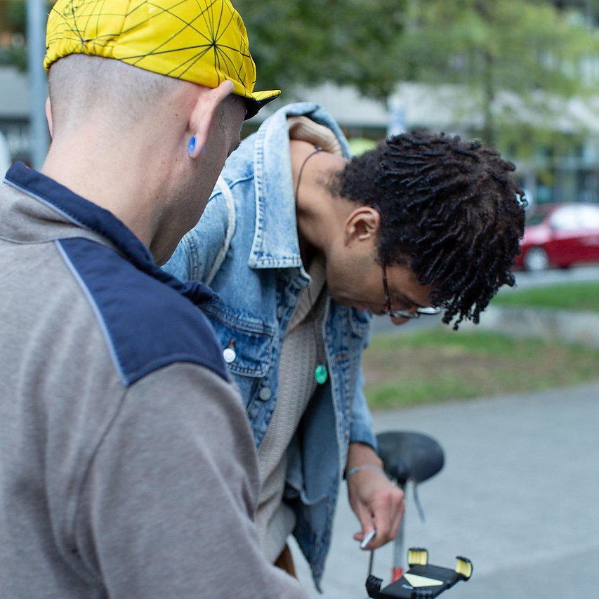 Hilfe zur Selbst-Hilfe von der Fahrrad-Reparatur-Community: zwei Männer beugen sich über ein Rad und reparieren die Halterung für das Smartphone am Lenker.