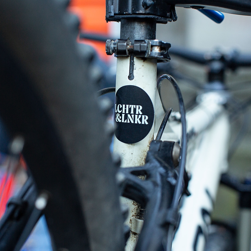 Ein Rad fotografiert von hinten: an der Lenker-Stange unter dem Sattel klebt ein neuer Aufkleber mit dem Logo der Fahrrad-Reparatur-Community LCHTR&LNKR (gesprochen Lichter & Lenker)