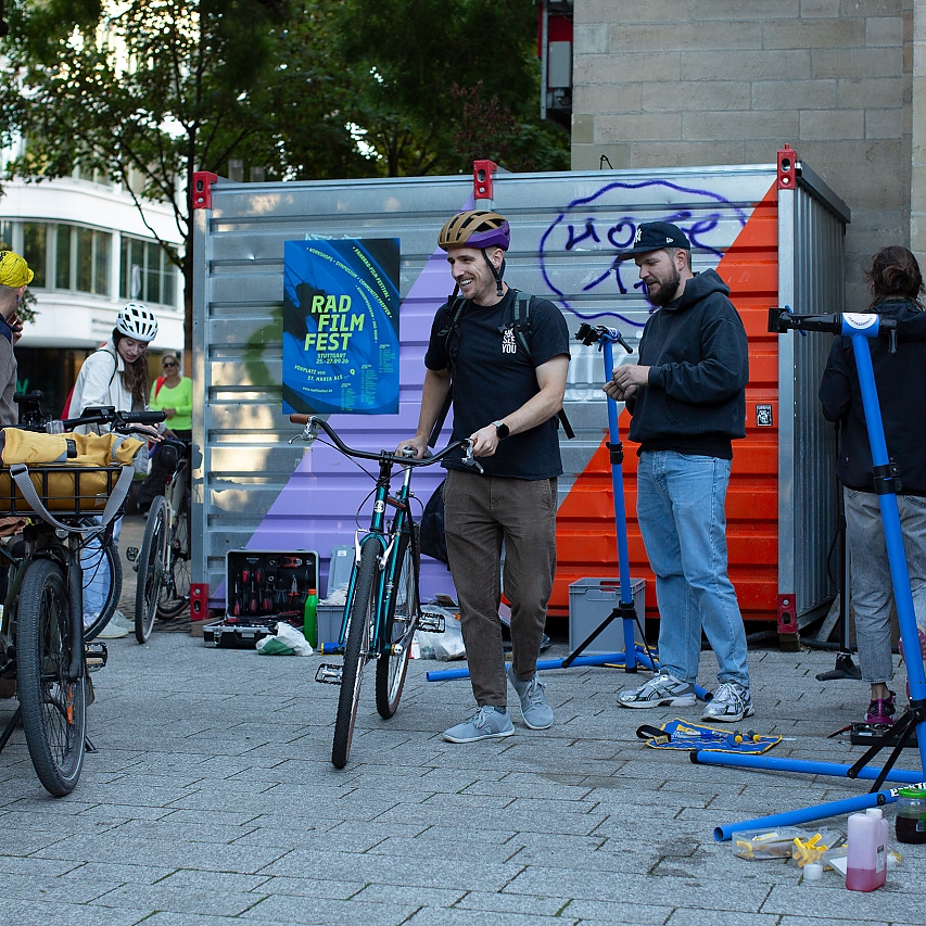 Glücklich verlässt ein Radfahrer mit seinem Rad die kostenfreie Fahrrad-Werkstatt von Licht & Lenker. Und schon die nächsten Kund:innen stehen da. In der Szene ebenso mit im Bild der Handwerker und sein Zubehör sowie Werkzeug. Daneben stehen ferner ein Trekking- und ein Lasten-Rad.