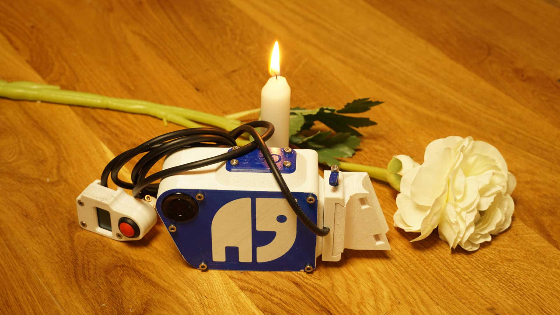 Holz-Tisch mit einer brennenden Kerze und einer weißen Rose. Davor liegt ein Gerät zur Abstands-Messung vom Projekt OpenBikeSensor. Es ist das persönliche Gerät von Andreas Mandalka aka Natenom und trägt daher sein Logo.