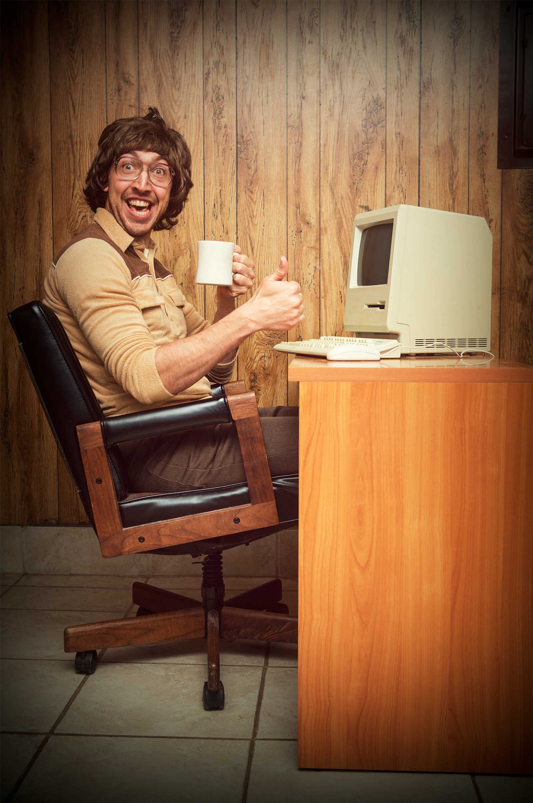 A vintage retro styled image with brown tones of a man in a wood paneled 1980s room sitting at his computer desk with a coffee cup in hand smiling. Bild: copy madiko / Foto: sjharmon